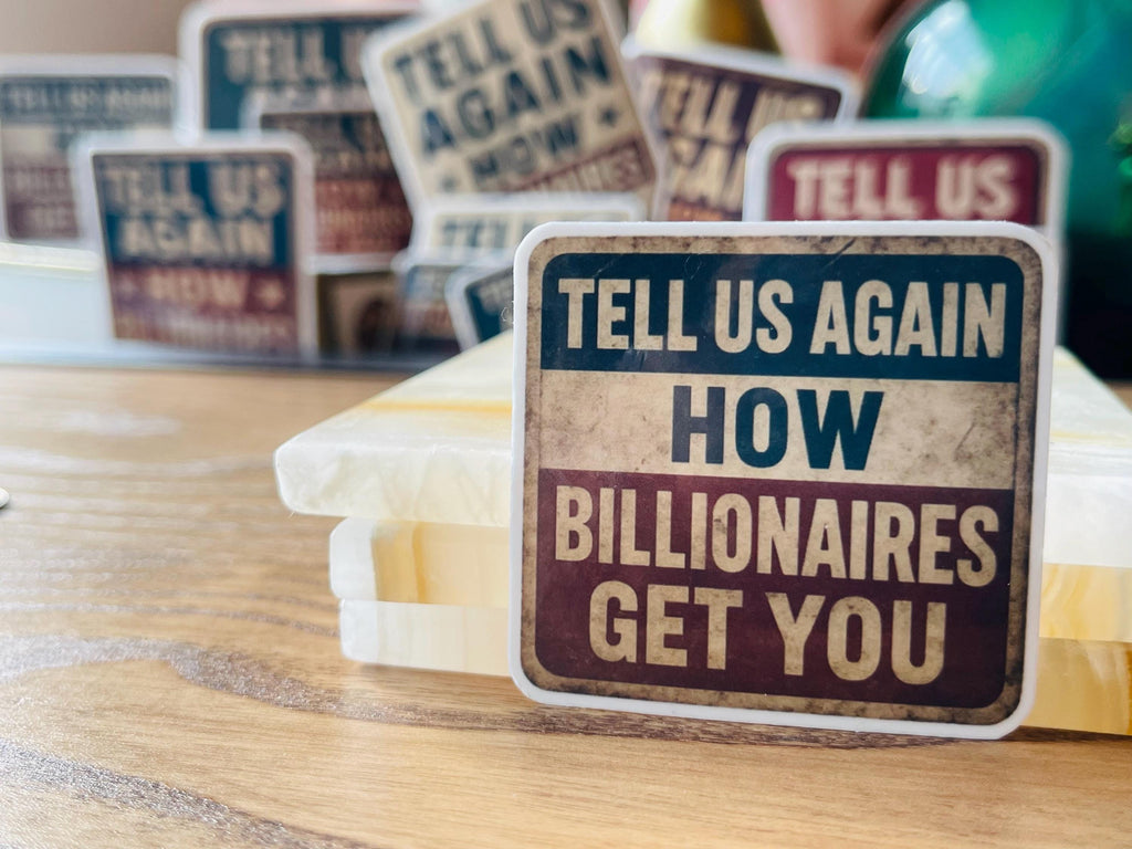 a close-up of a sticker with the text "TELL US AGAIN HOW BILLIONAIRES GET YOU" on a wooden surface.