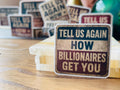 a close-up of a sticker with the text "TELL US AGAIN HOW BILLIONAIRES GET YOU" on a wooden surface.