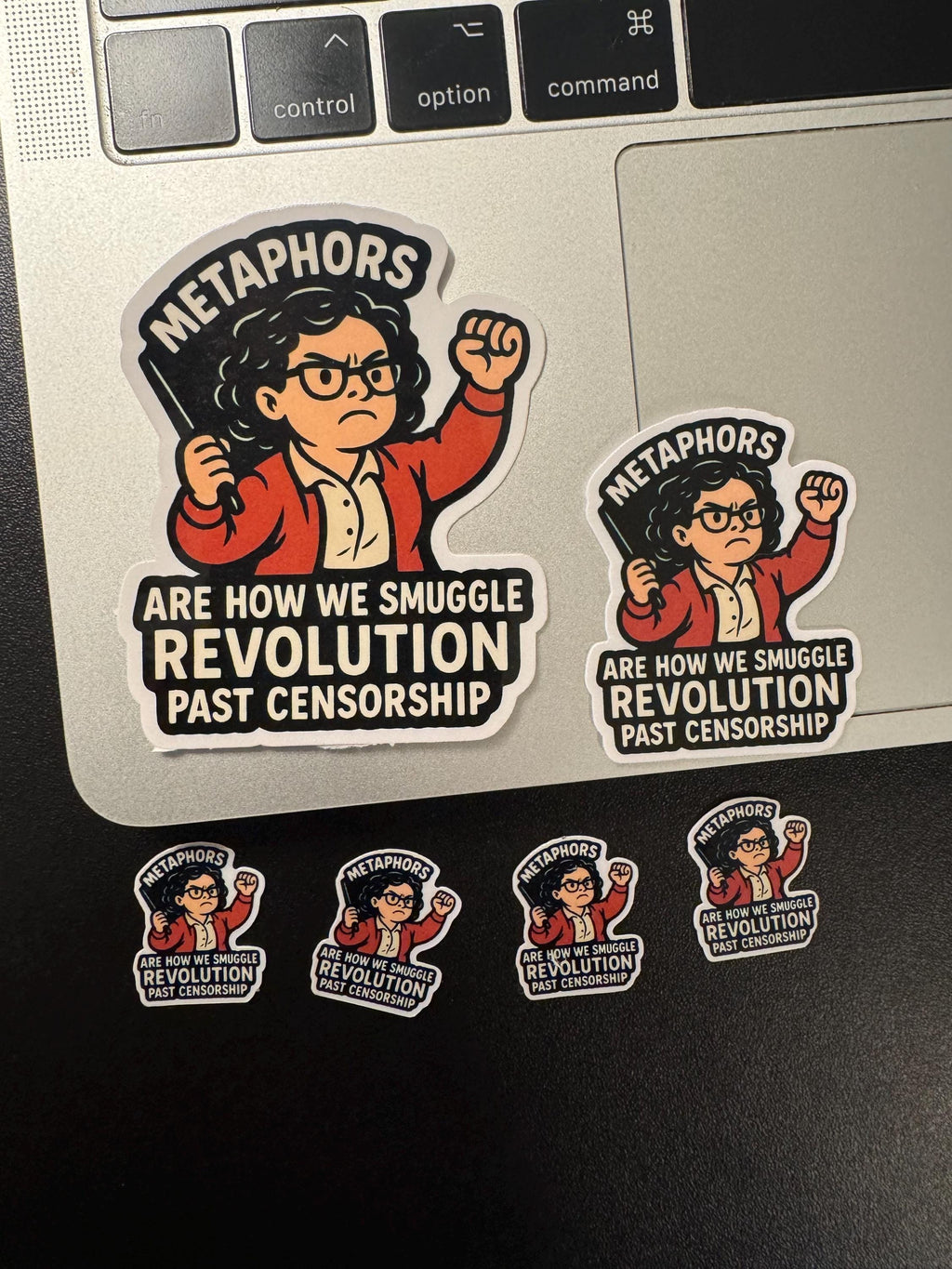 a sticker with a cartoon character and text, along with four smaller stickers below it. The larger sticker features a woman with a fist and the text "METAPHORS ARE HOW WE SMUGGLING REVOLUTION PAST CENSORSHIP". The smaller stickers also feature the same text and the same cartoon character.
