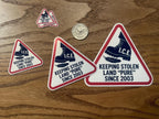 Five triangular abolish ice sticker designs in different sizes laid on a wood table next to a quarter for scale, each showing the I.C.E. boot stomping a feather.