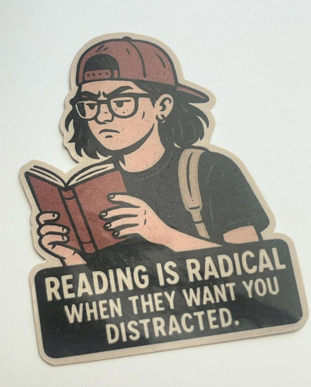 A sticker depicting a person reading a book, with the text "Reading is Radical When They Want You Distracted."