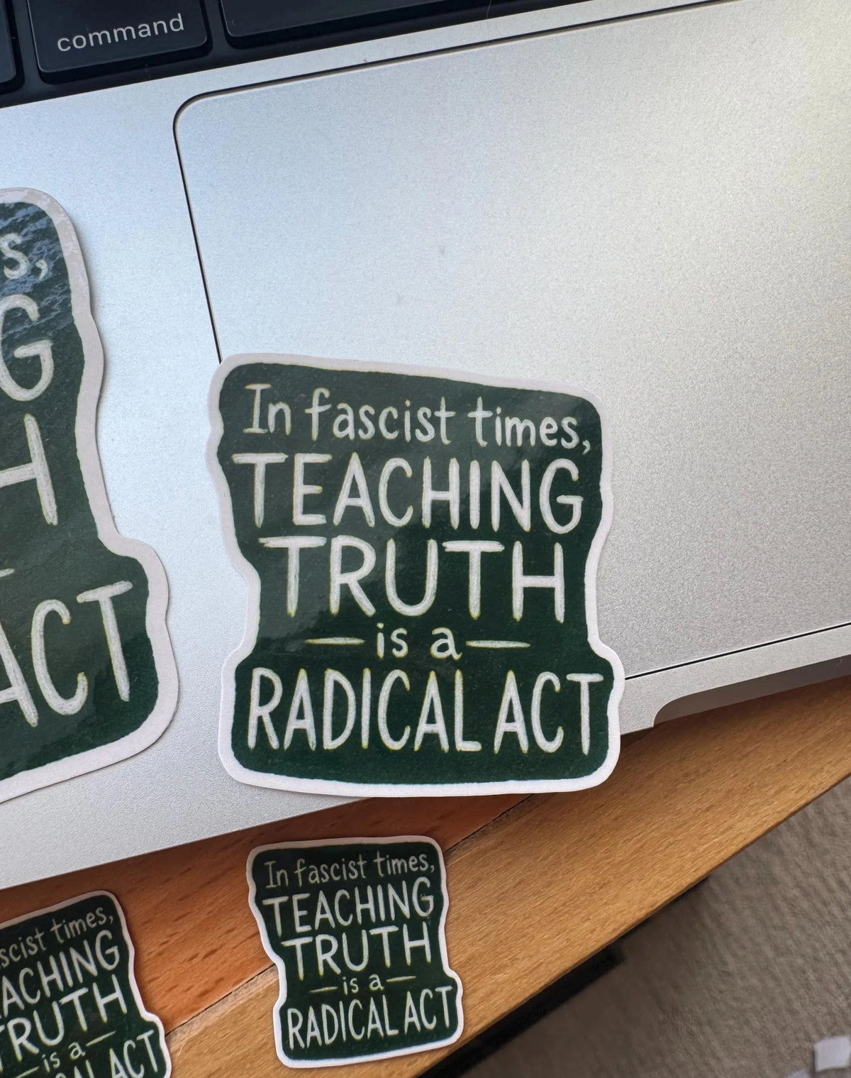 Close-up of an anti-fascist sticker reading 'In fascist times, teaching truth is a radical act' placed near the corner of a laptop, with partial views of matching stickers on the wooden desk surface around it.
