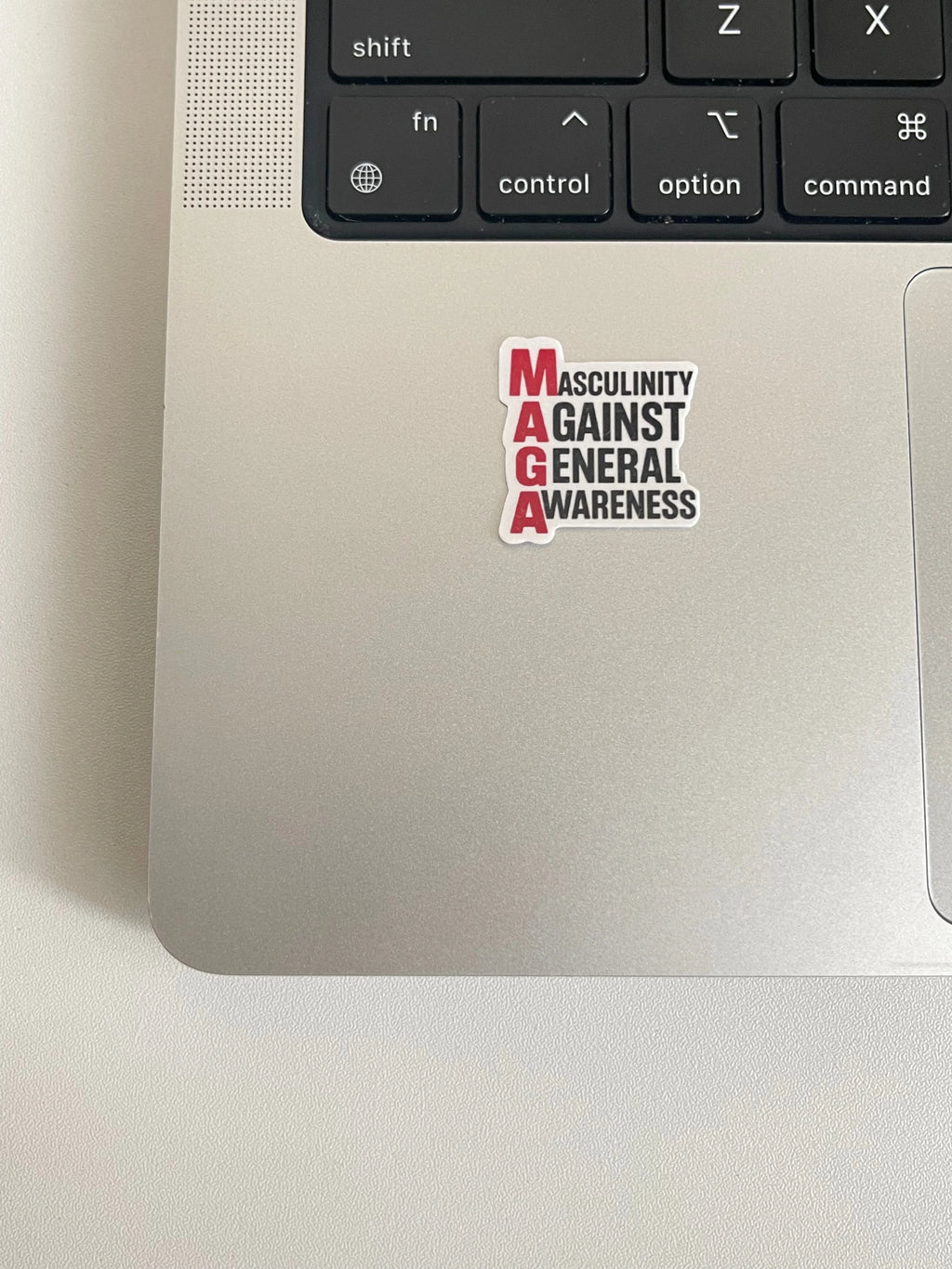 A single small Anti Trump 'Masculinity Against General Awareness' sticker placed on the silver trackpad area of a laptop