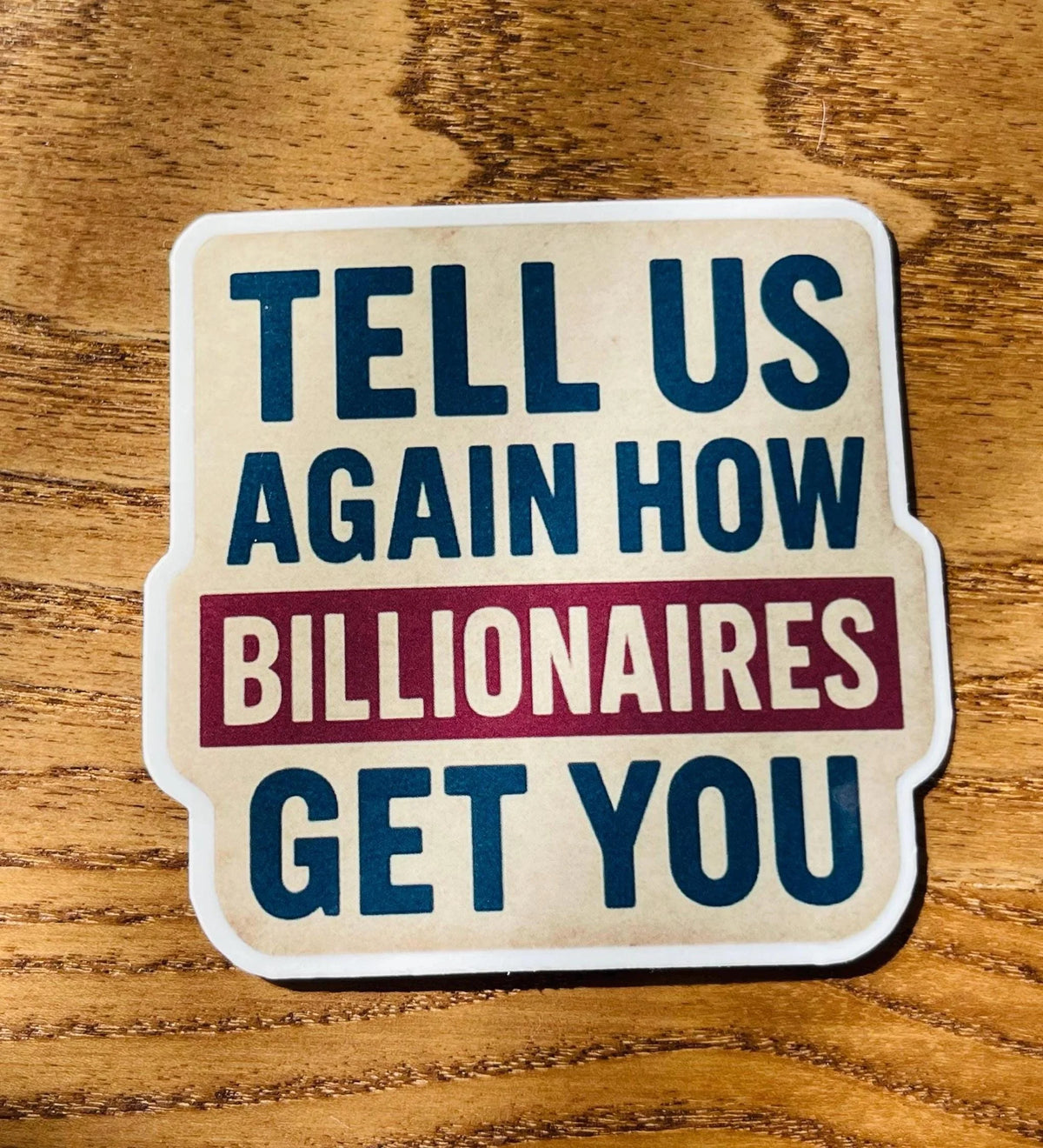 Anti Trump satire sticker with cream background and scalloped white border resting on a wood-grain tabletop, displaying the full "Tell Us Again How Billionaires Get You" text.