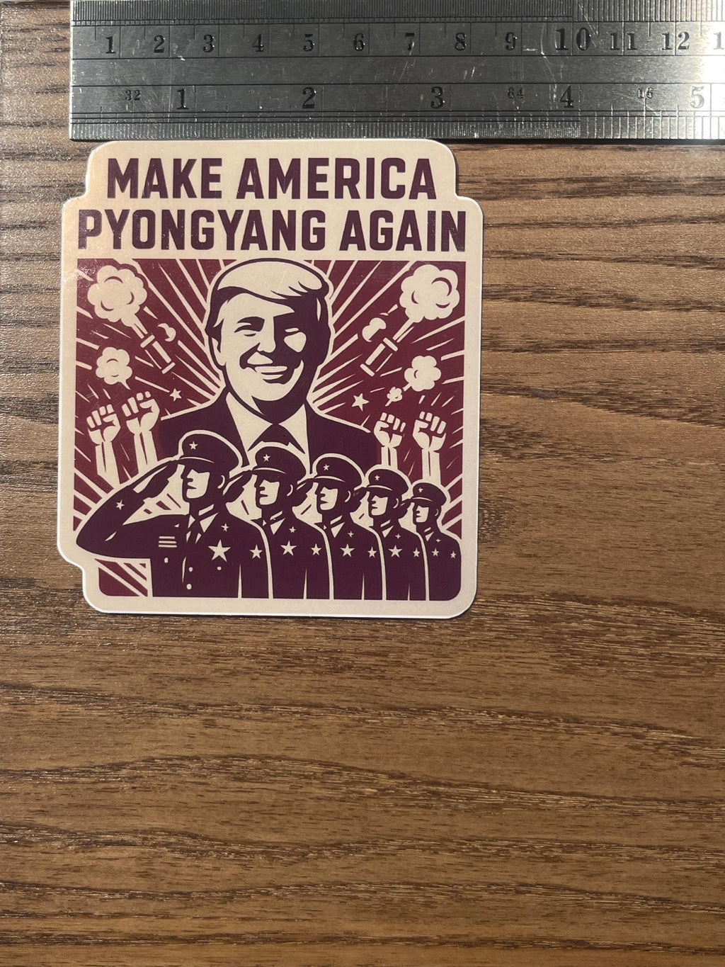 Anti-fascist sticker measured against a metal ruler on a wood surface, showing the 'Make America Pyongyang Again' propaganda-style parody design with a Trump portrait, sunburst rays, smoke bursts, raised fists, and a row of saluting uniformed figures.