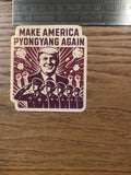 Anti-fascist sticker measured against a metal ruler on a wood surface, showing the 'Make America Pyongyang Again' propaganda-style parody design with a Trump portrait, sunburst rays, smoke bursts, raised fists, and a row of saluting uniformed figures.