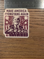 Anti-fascist sticker measured against a metal ruler on a wood surface, showing the 'Make America Pyongyang Again' propaganda-style parody design with a Trump portrait, sunburst rays, smoke bursts, raised fists, and a row of saluting uniformed figures.