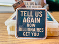 A sticker with the text "TEELL US AGAIN HOW BILLIONAIRES GET YOU" on a wooden surface.