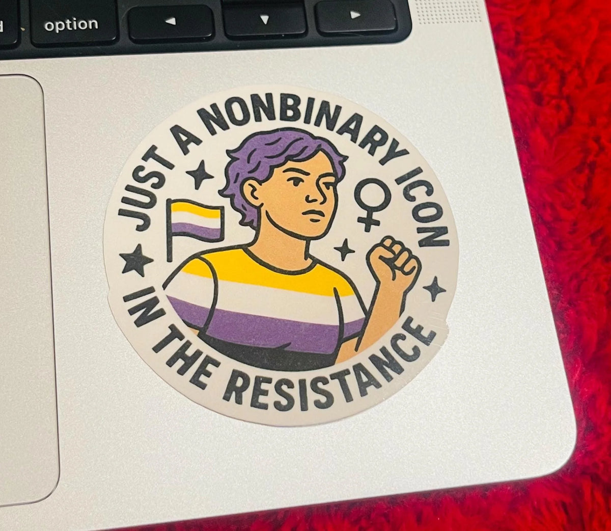 Circular nonbinary pride design stuck near the top corner of a laptop keyboard, showing the illustrated figure with raised fist and the text 'Just a Nonbinary Icon in the Resistance,' photographed as a funny laptop sticker beside a red furry surface.