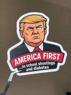 FDT-themed sticker showing a scowling Trump caricature and 'America First...in school shootings and diabetes' text, held up with tweezers against a plain brown wall.