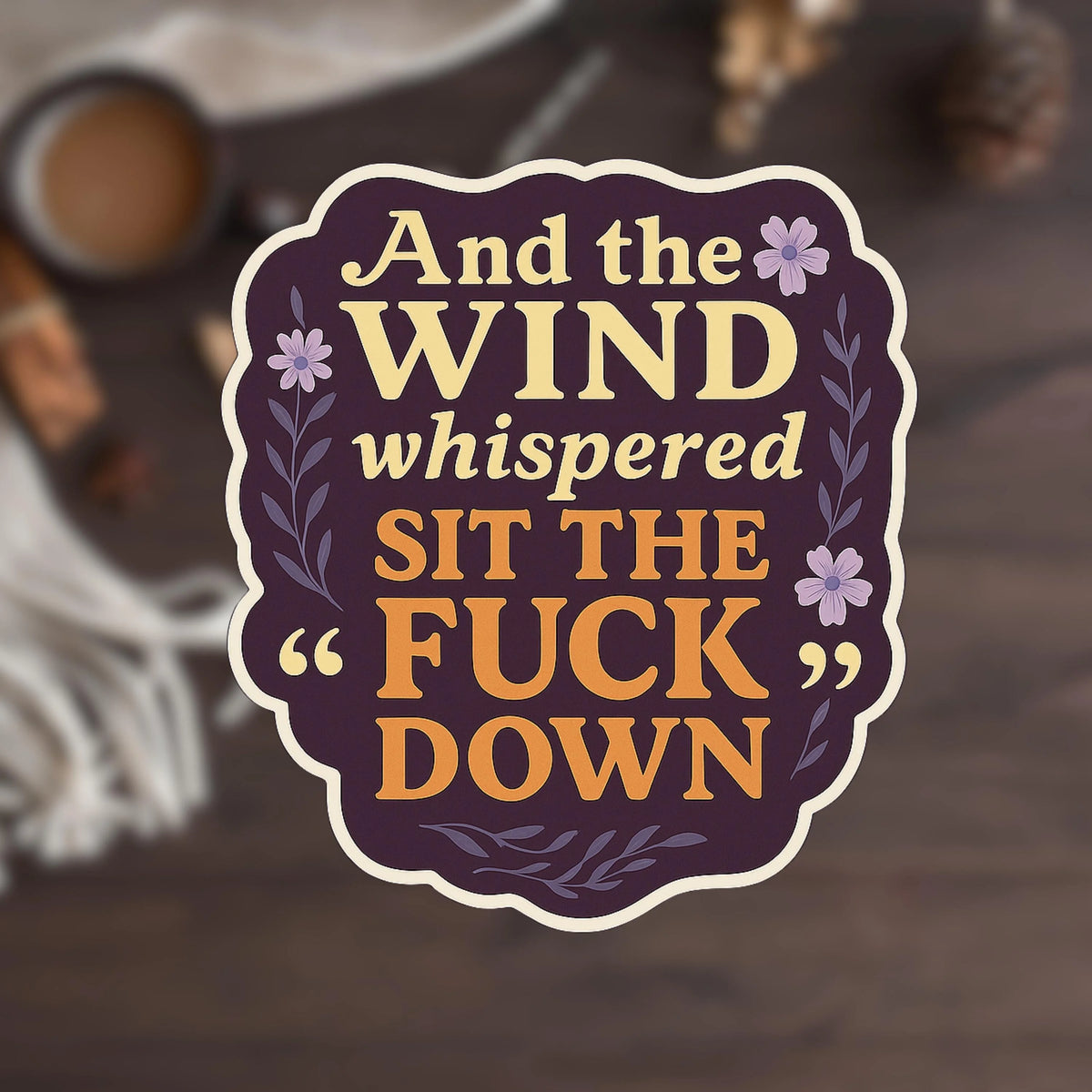 Funny floral sticker in dark plum tones with floral accents reading 'And the Wind whispered SIT THE FUCK DOWN,' shown against a softly blurred coffee mug and wooden chair background.