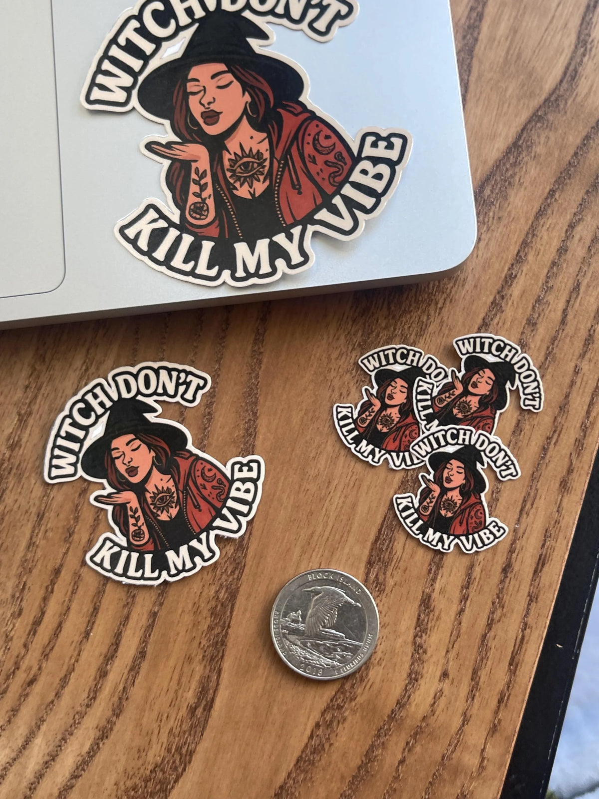 Halloween sticker of a witch blowing a kiss applied to the corner of a laptop, with several smaller matching stickers and a quarter for size scale on a wood surface.