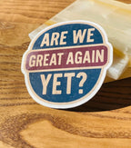 Circular liberal sticker with the text 'Are We Great Again Yet?' in navy and maroon tones, resting on a wood table next to its clear plastic packaging sleeve.