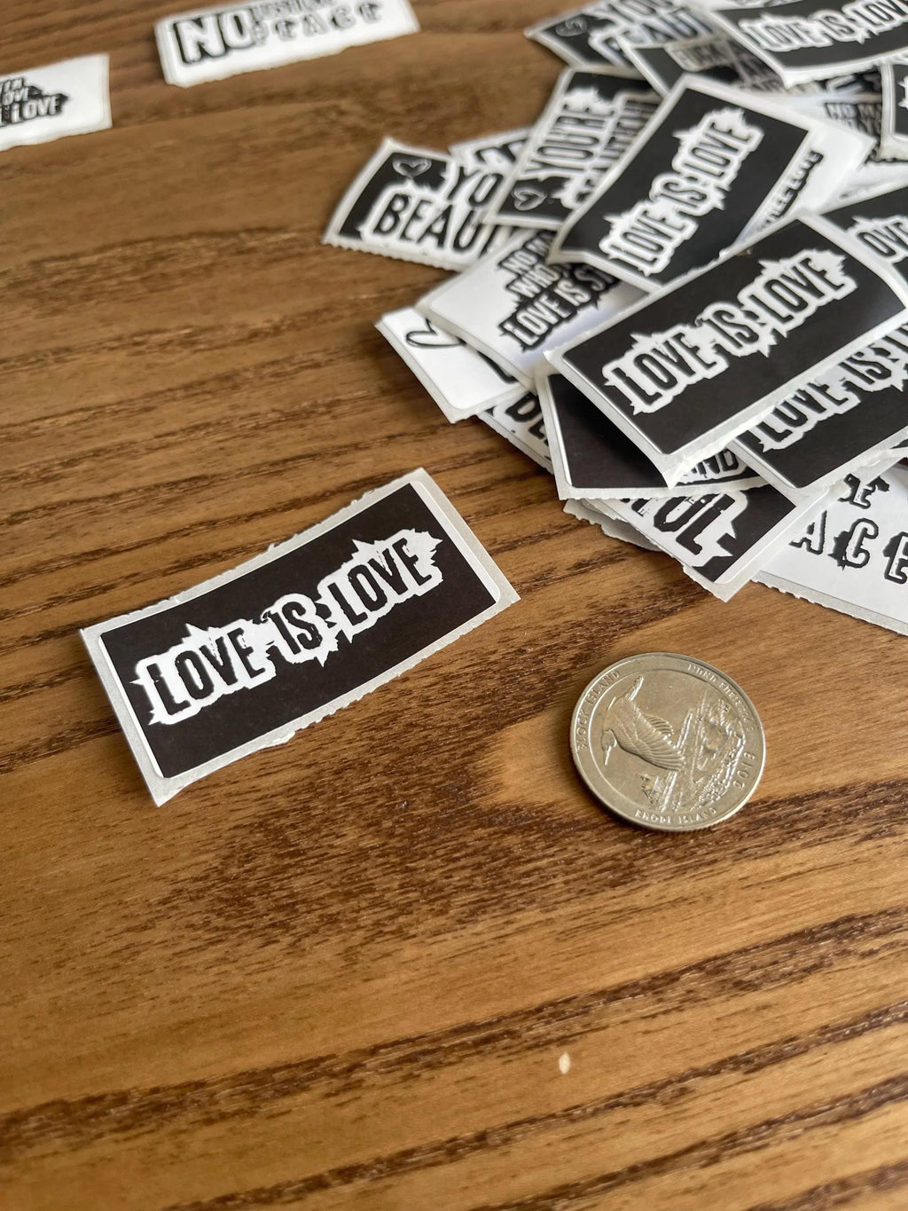 A Love Is Love sticker placed on a wooden surface near a quarter for size reference, with a pile of other slogan stickers scattered in the background.