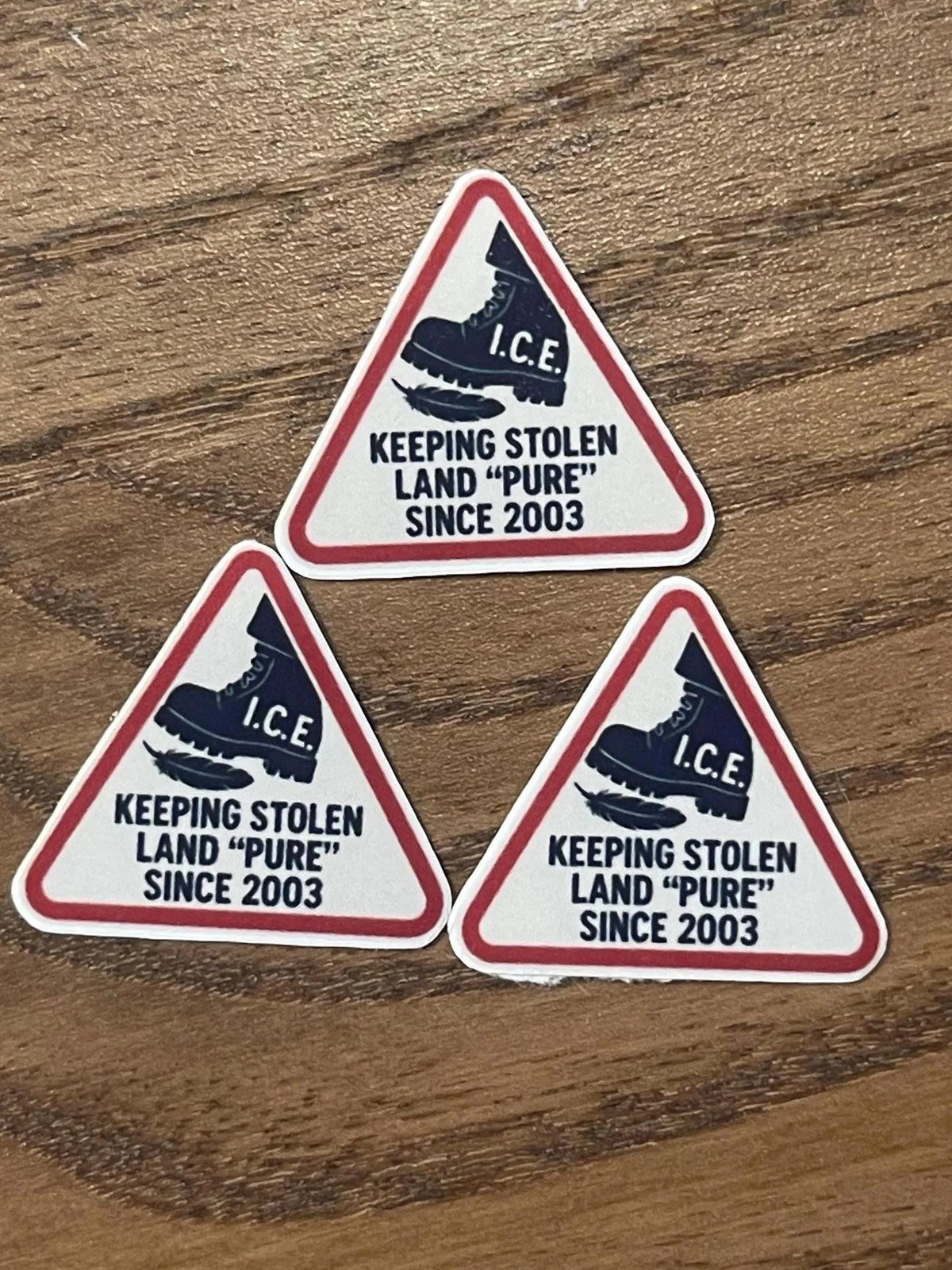 Three triangular sticker meme designs featuring the I.C.E. boot stomping a feather arranged on a wood table.
