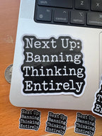 Resist sticker reading 'Next Up: Banning Thinking Entirely' stuck on a laptop lid next to a quarter coin for size reference, with smaller matching pieces nearby.