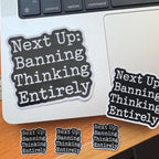Liberal sticker set showing large and small versions of 'Next Up: Banning Thinking Entirely' applied to a laptop lid and scattered across a wooden desk.
