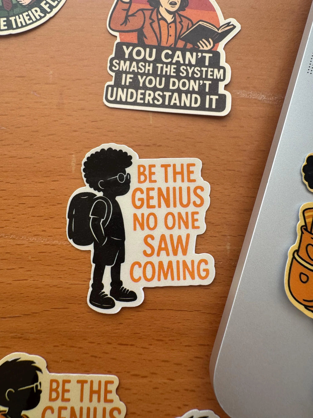 Student sticker featuring a silhouetted Black boy with an afro and backpack next to orange 'Be The Genius No One Saw Coming' text, resting on a wooden desk among other illustrated stickers.