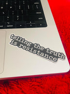"Telling the truth is resistance" vinyl decal applied to the corner of a silver laptop trackpad area near the keyboard, resting on a red fuzzy surface.
