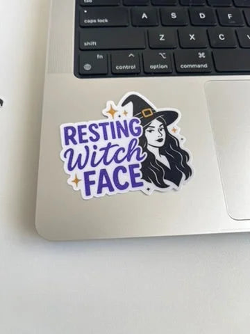 Purple and black 'Resting Witch Face' witch sticker with a witch hat design, shown on the corner of a silver laptop near the keyboard